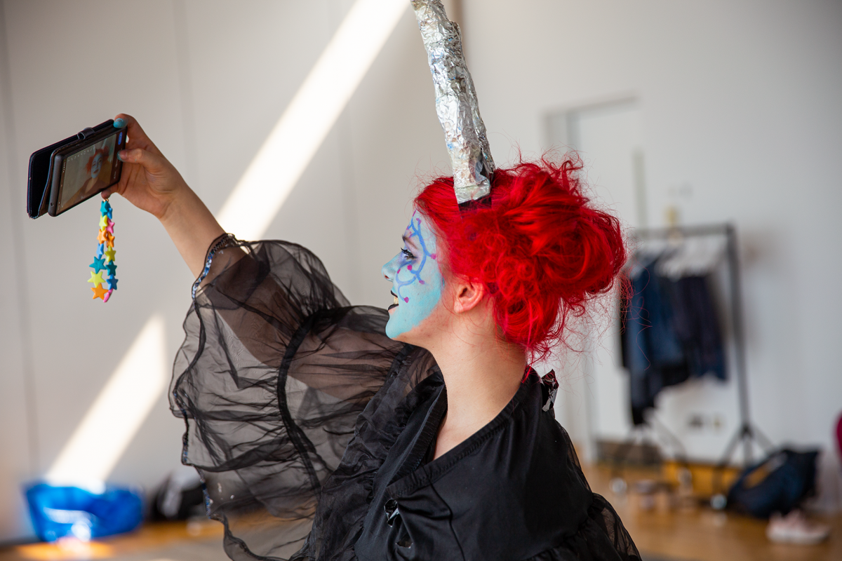 Work Title: Amplify development weekend costume rehearsal Project: Amplify Photo Credit: Josie Young A young performer in full costume with bright hair and a head piece holding their phone above their head taking a selfie