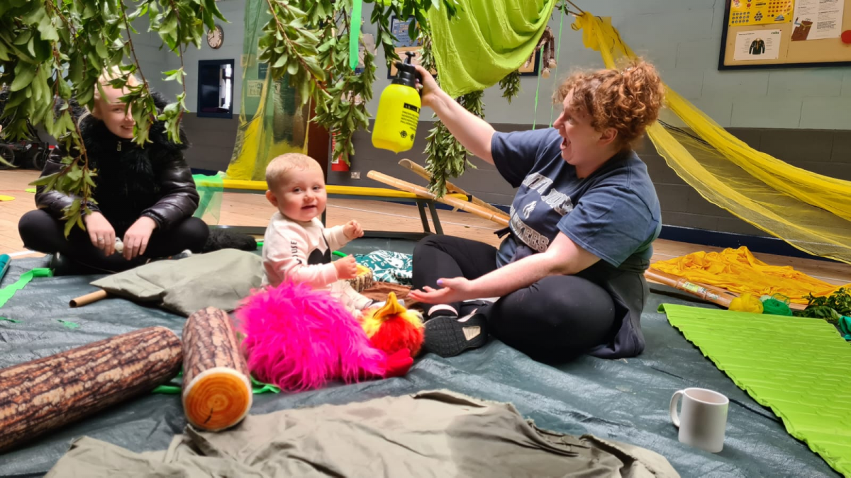 Young baby sits in a jungle inspired room, whilst artist Roz McAndrews sprays water over her. Child’s grown-up looks at them smiling